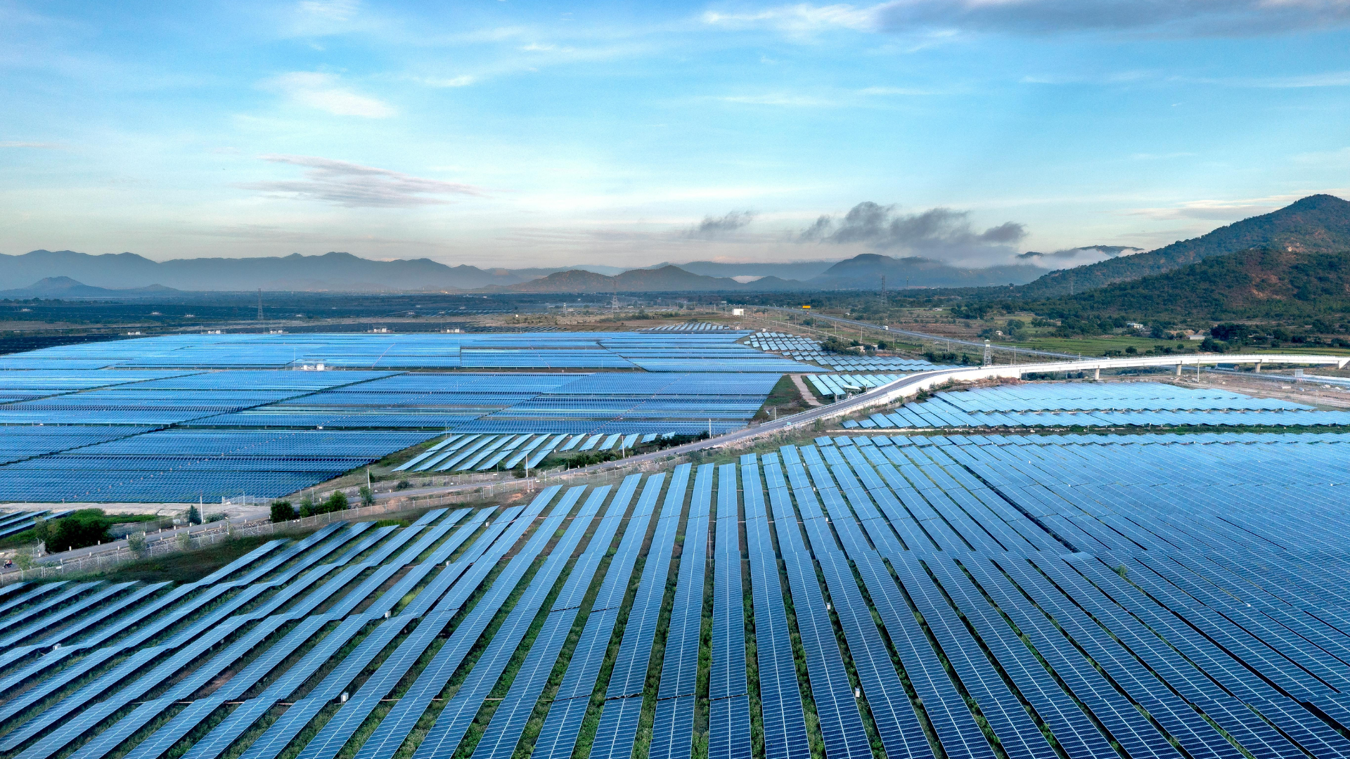Aerial view of a vast solar farm with rows of blue solar panels and distant mountains in the background on a clear day.