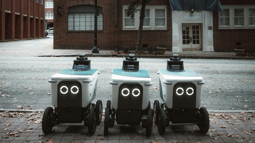 Three delivery robots on wheels with glowing LED eyes parked on a city street, front of a brick building and fallen leaves around them.