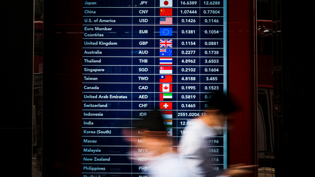 Large vertical display of a currency exchange board showing country names, currency codes, flags, and exchange rates; blurred pedestrians move in front of it.