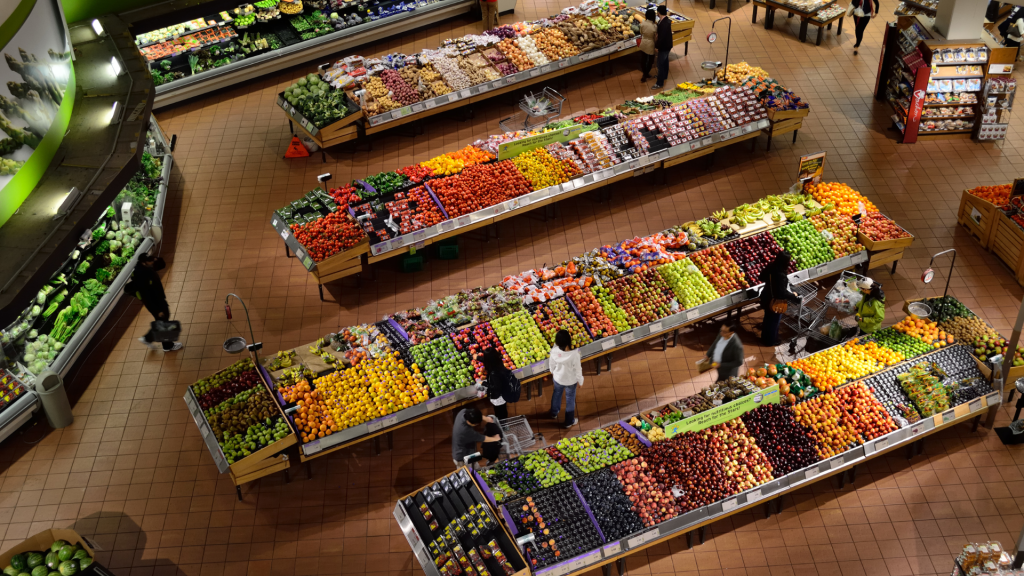 Aerial/top-down view of a supermarket produce department with long rows of colorful fruits and vegetables and shoppers browsing.