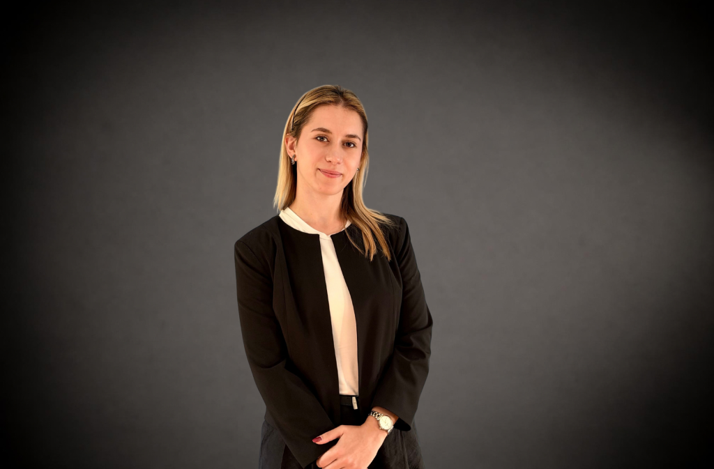 Professional woman in a black blazer and white blouse, arms loosely crossed in front, posed against a dark gradient background.