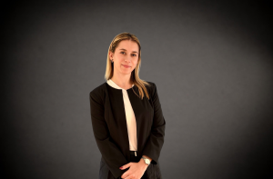 Professional woman in a black blazer and white blouse, arms loosely crossed in front, posed against a dark gradient background.