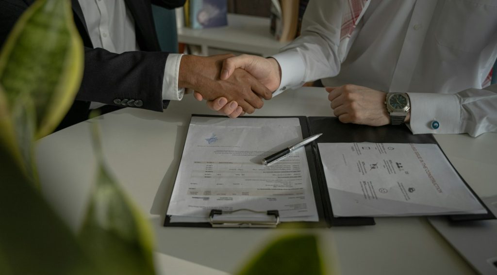 Two professionals shake hands over a signed contract on a clipboard with a pen and documents on the table behind them.